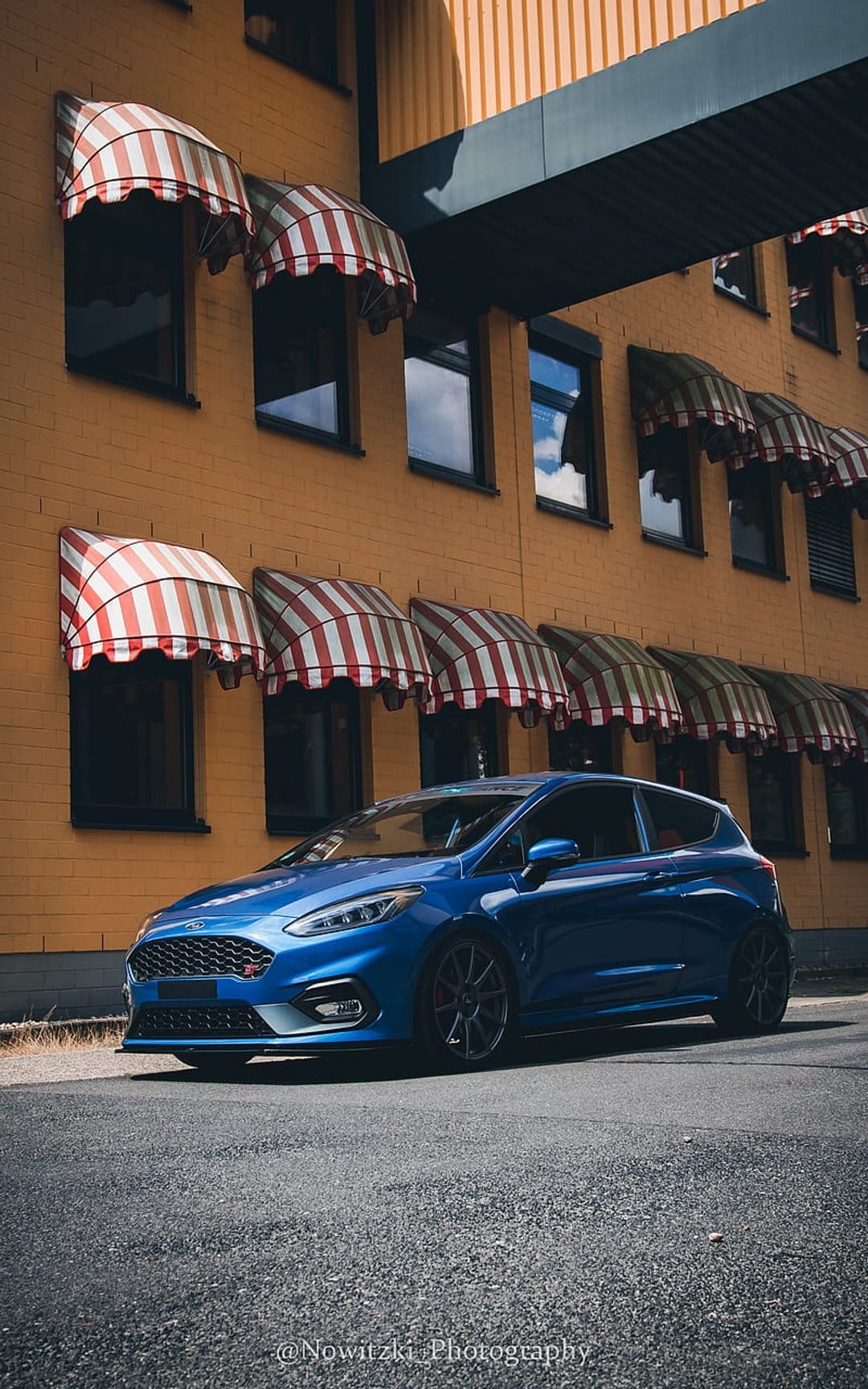 Blue Ford Fiesta ST hatchback parked on asphalt in front of a yellow building with striped window awnings.