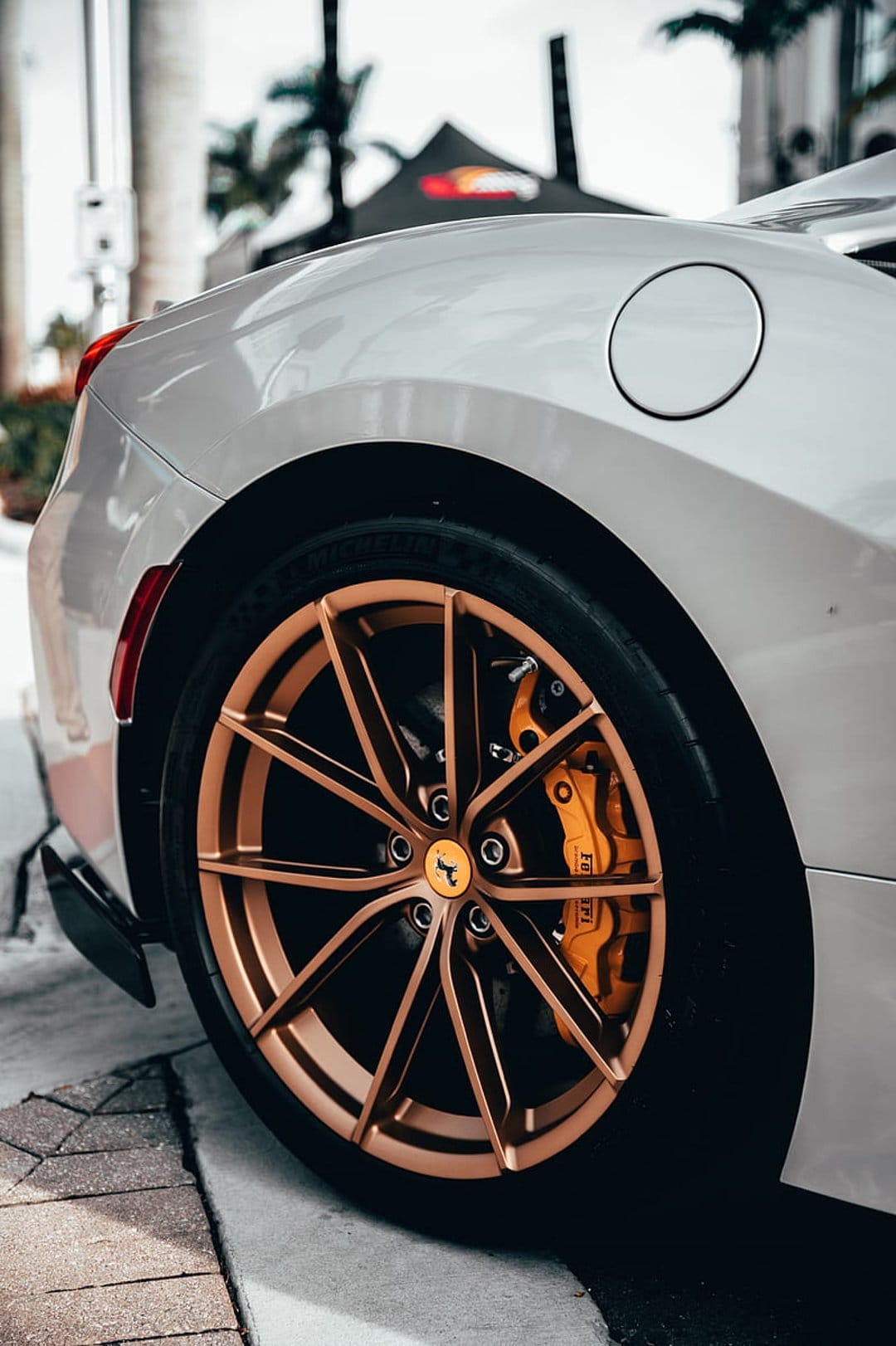 White Ferrari rear wheel featuring bronze multi-spoke alloy rims and bright orange brake calipers against palm trees.