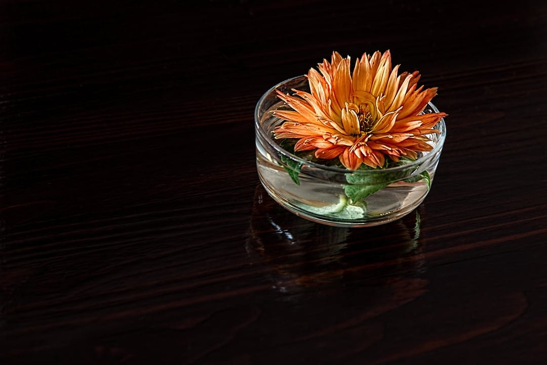 Orange chrysanthemum flower floating in a water-filled glass bowl on a dark reflective wooden table.