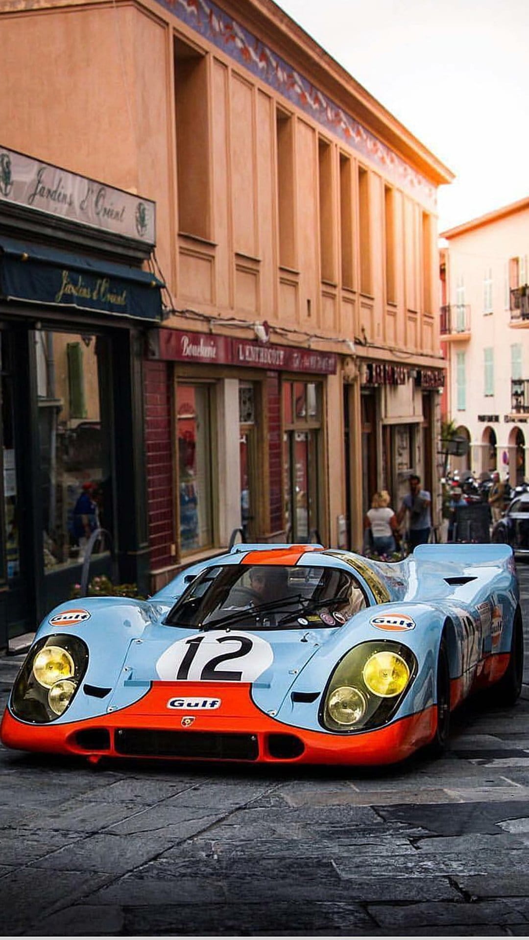 Porsche 917 race car with Gulf livery parked on a cobblestone street during golden hour in a European town.
