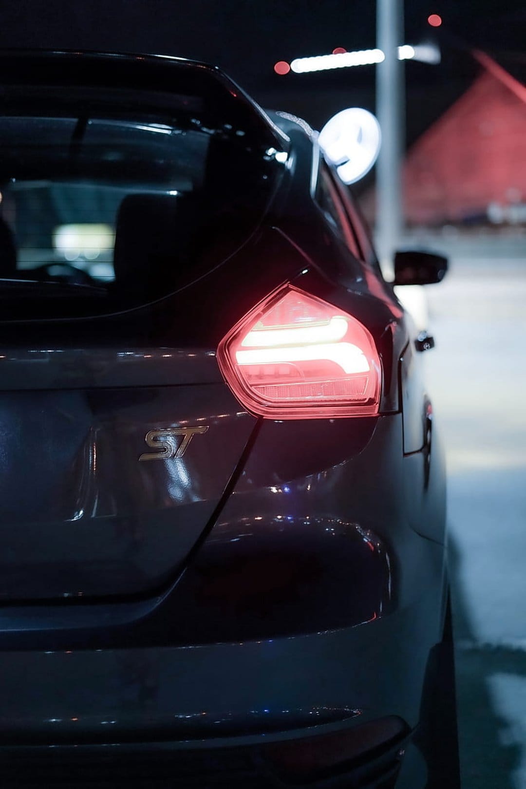 Red LED taillight of a black Ford Focus ST glows at night alongside a metallic silver ST badge in close-up.