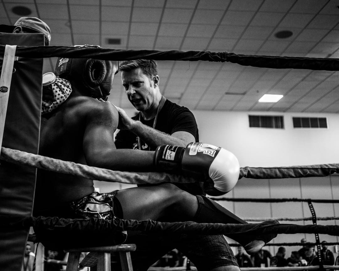 Muay Thai fighter in headgear sits on a stool while a coach adjusts his gloves in a black and white boxing ring.