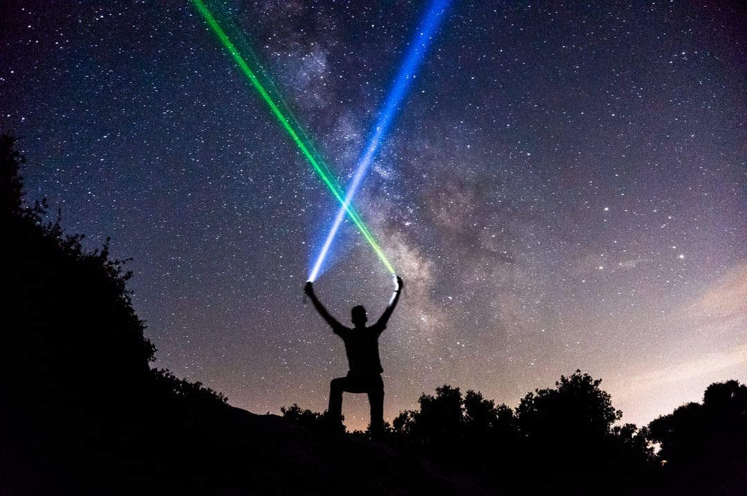 Person in silhouette crosses glowing blue and green light blades against a vivid Milky Way galaxy night sky.