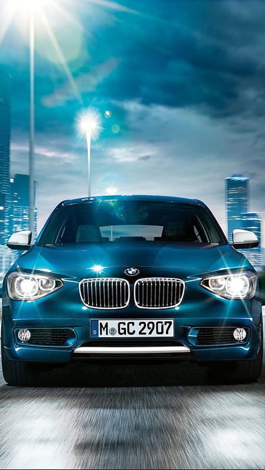 Teal BMW luxury sedan driving on an urban road at night with glowing headlights and a blurred city skyline backdrop.
