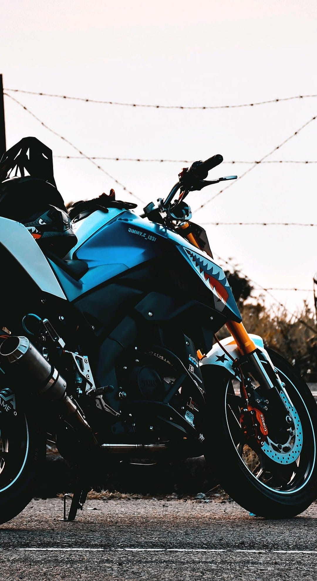 Blue motorcycle with a shark-mouth decal parked next to a barbed wire fence against a bright, hazy sky background.
