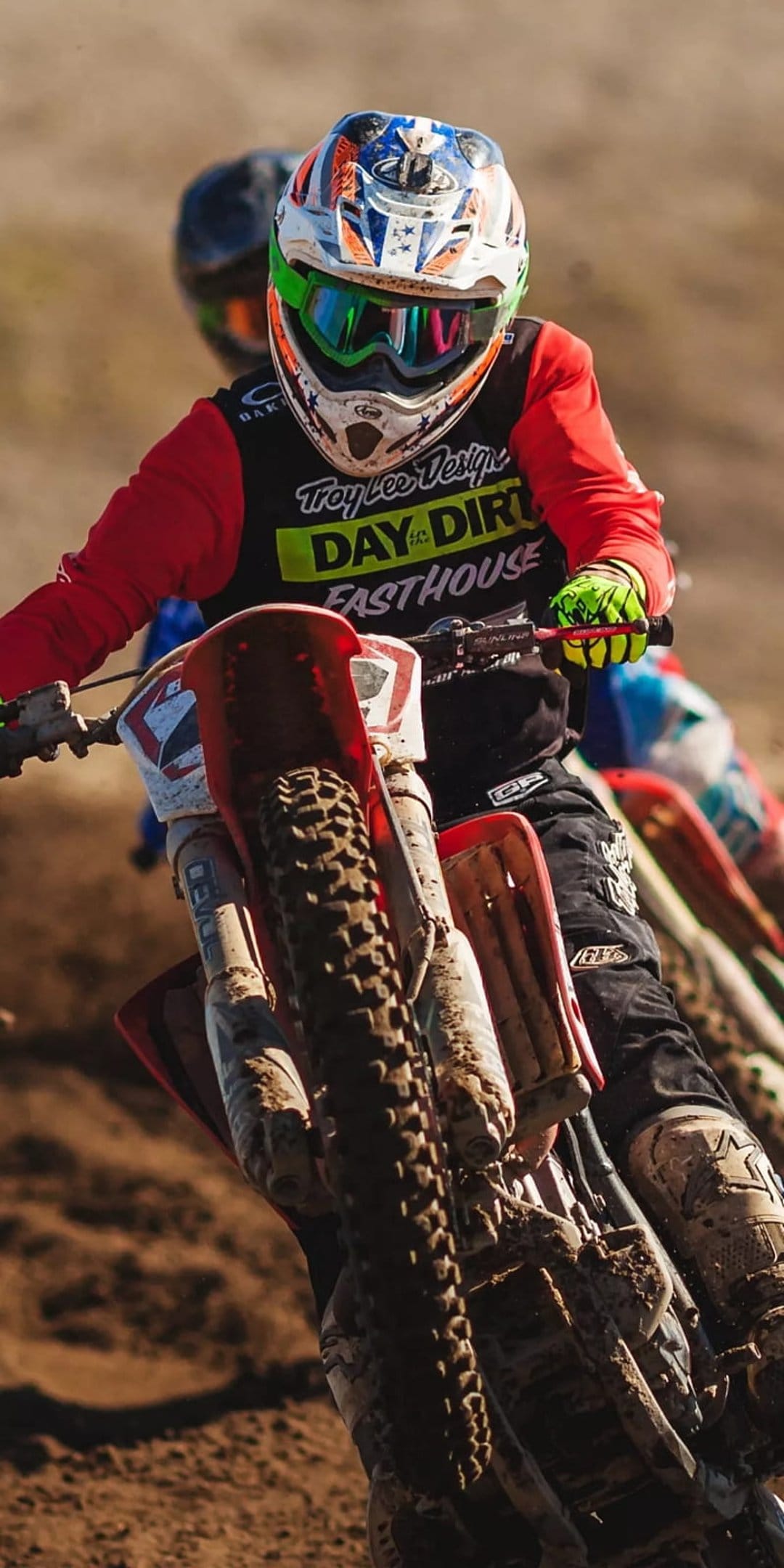 Motocross rider in a red jersey leans a dirt bike into a muddy corner with dirt splashing from the front tire.