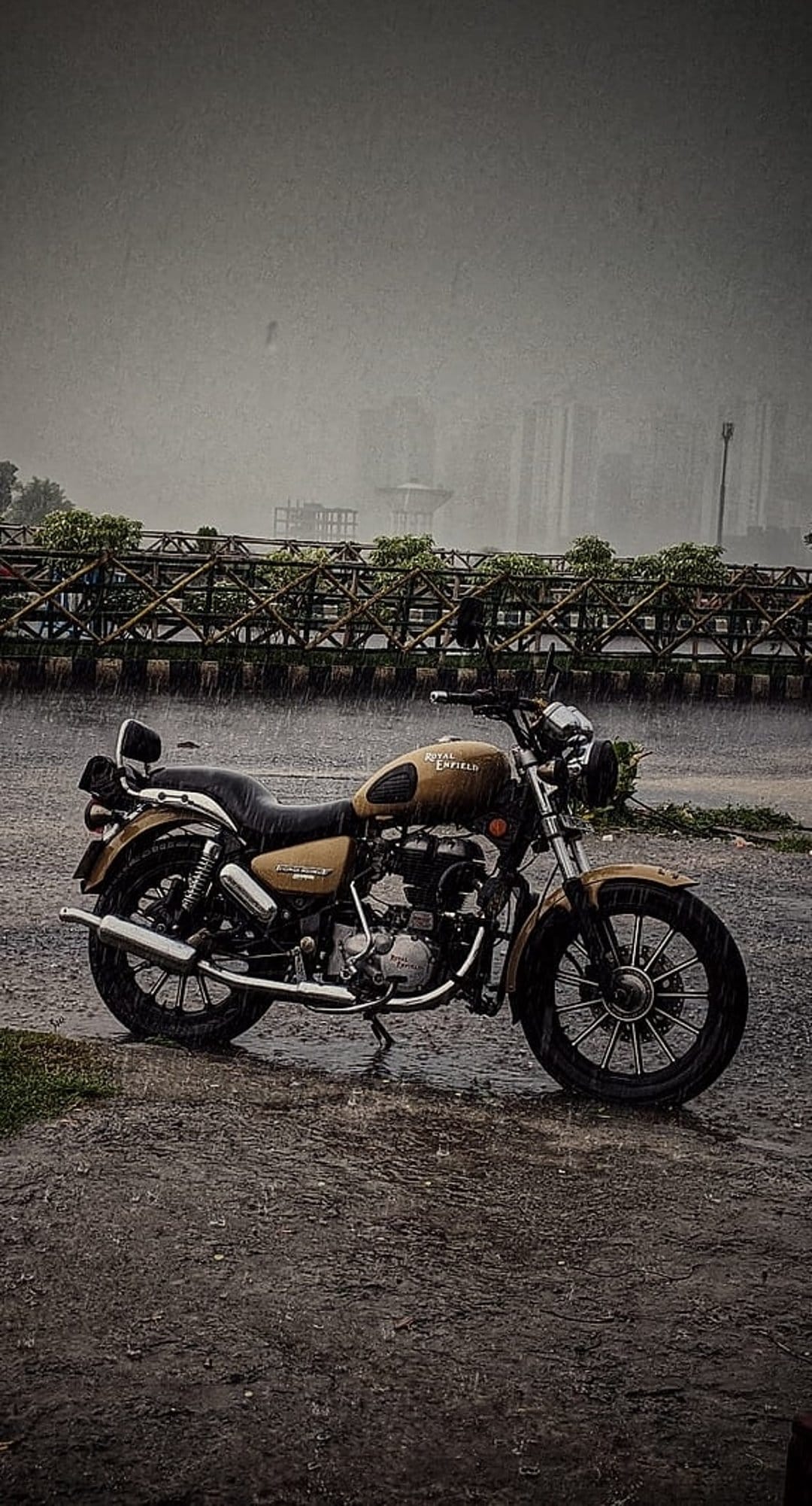 Royal Enfield motorcycle parked on a wet urban road during heavy rain with a wooden bridge in the background.