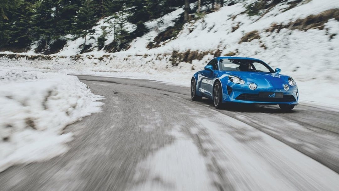 Blue Alpine A110 sports car driving on a winding snowy mountain road with evergreen trees in the background.