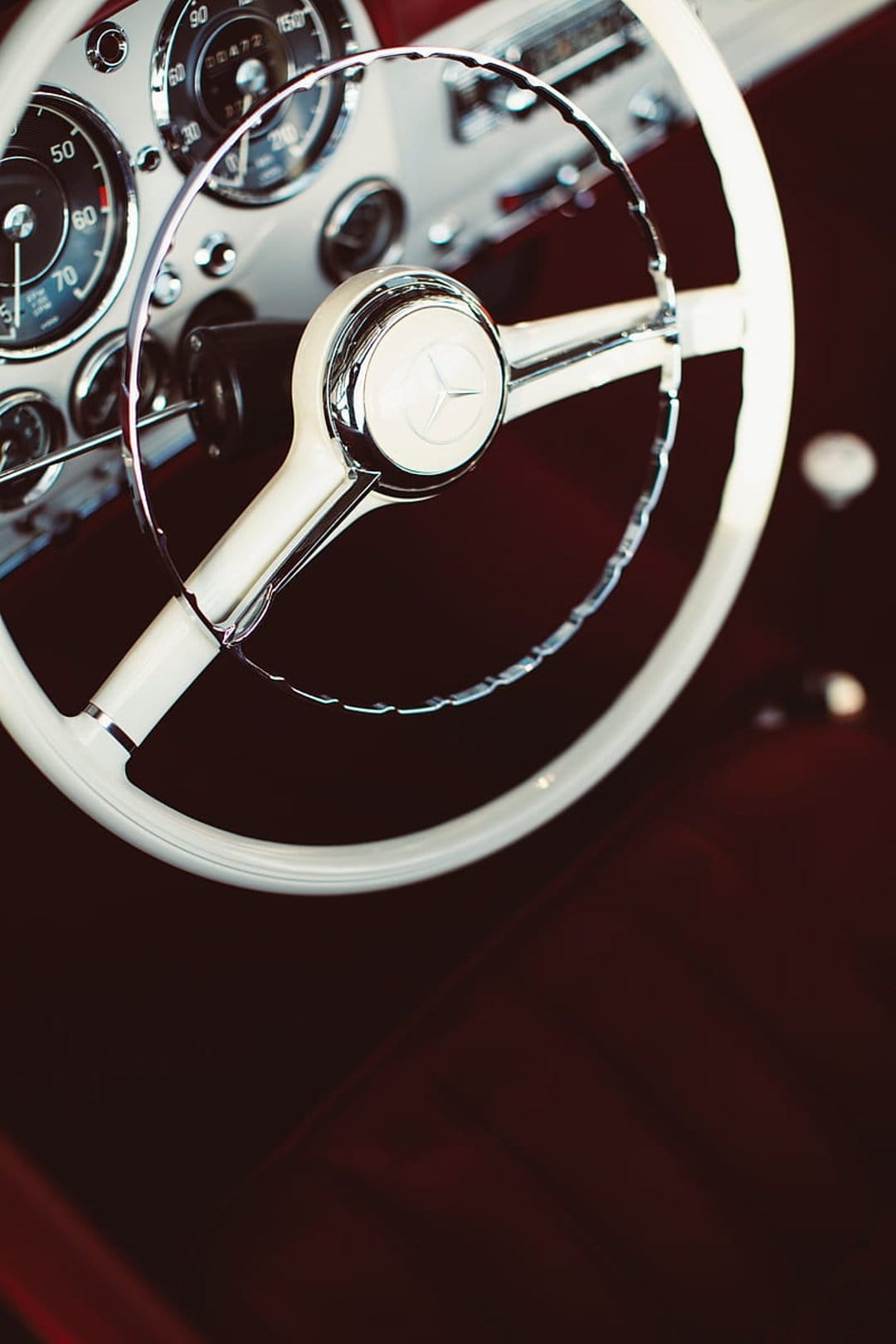 White Mercedes-Benz steering wheel with a chrome horn ring set against a red leather dashboard and analog gauges.