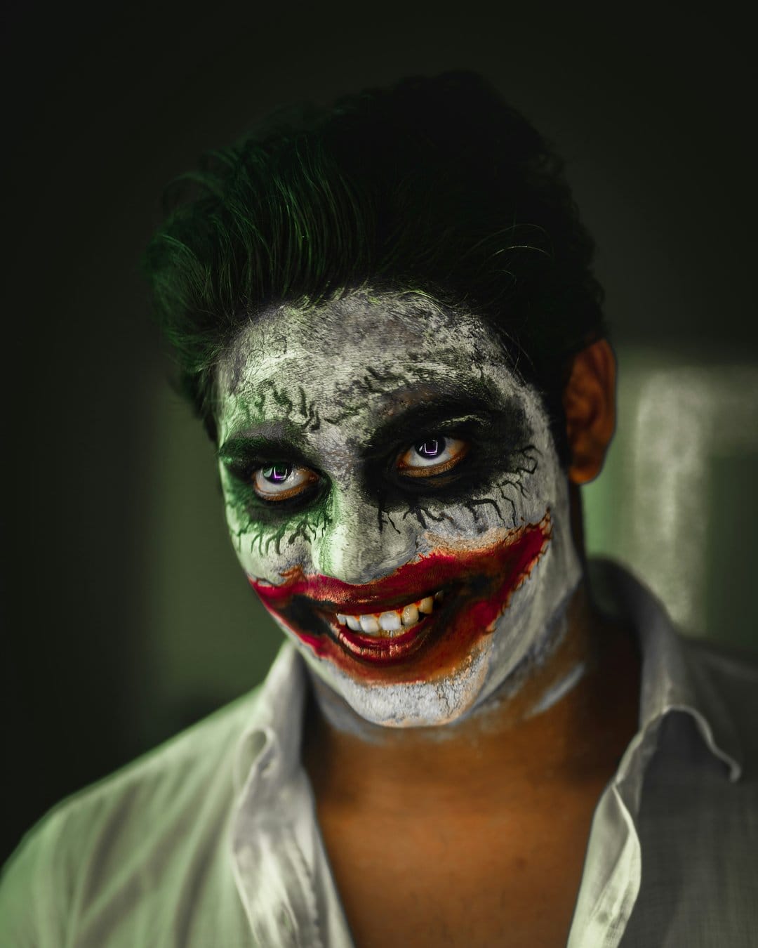 Man wearing cracked white clown face paint with dark eyes and a red smile in a moody vertical portrait.