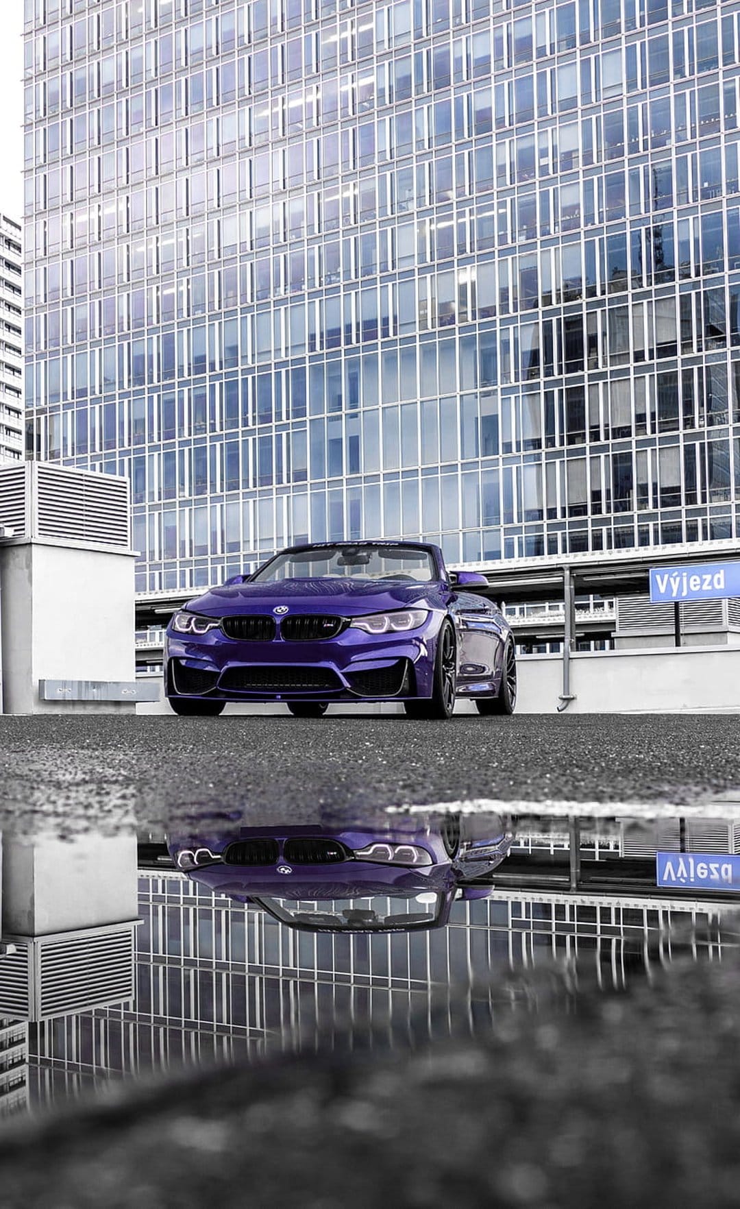 Purple BMW M4 convertible parked on wet asphalt reflects in rainwater near a glass urban building at night.