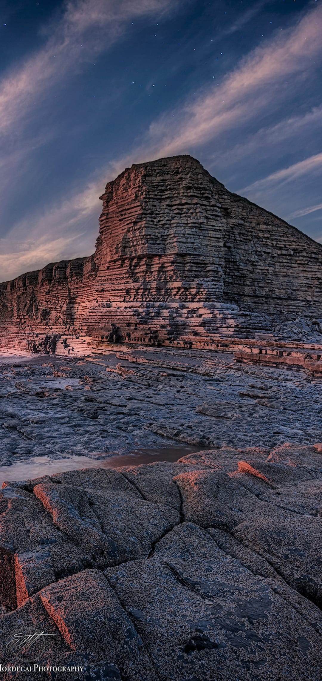 Stratified rock cliff rises toward a starry twilight sky with orange horizon glows above a rugged coast.
