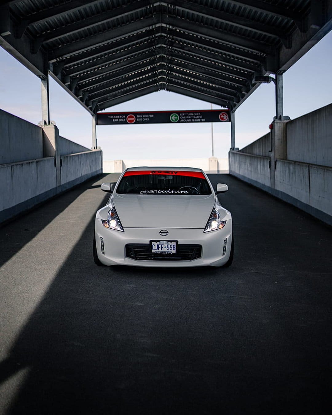 White Nissan 370Z with a red windshield banner driving under a concrete walkway with its headlights turned on.