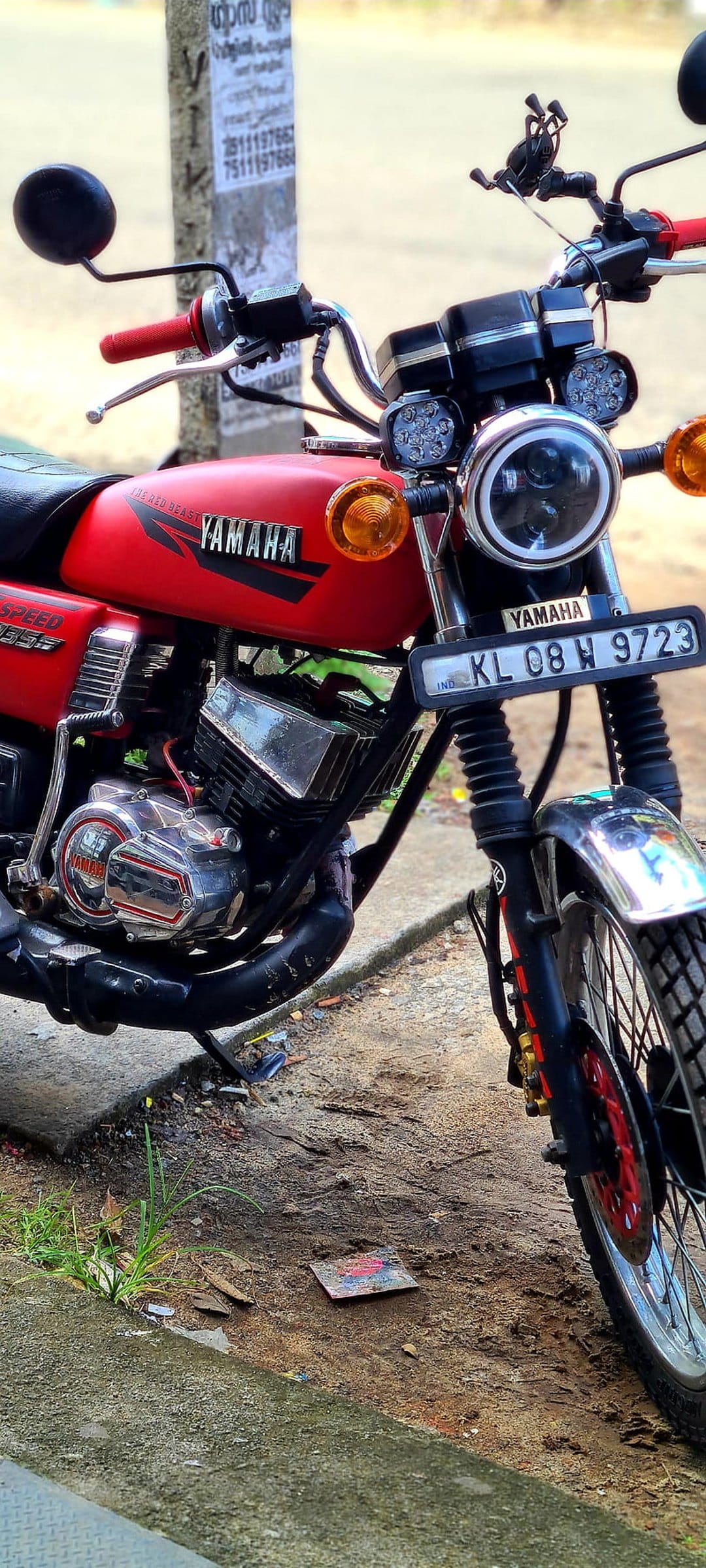 Red Yamaha RX 100 motorcycle with a round projector headlamp and custom LED lights in a vertical view.