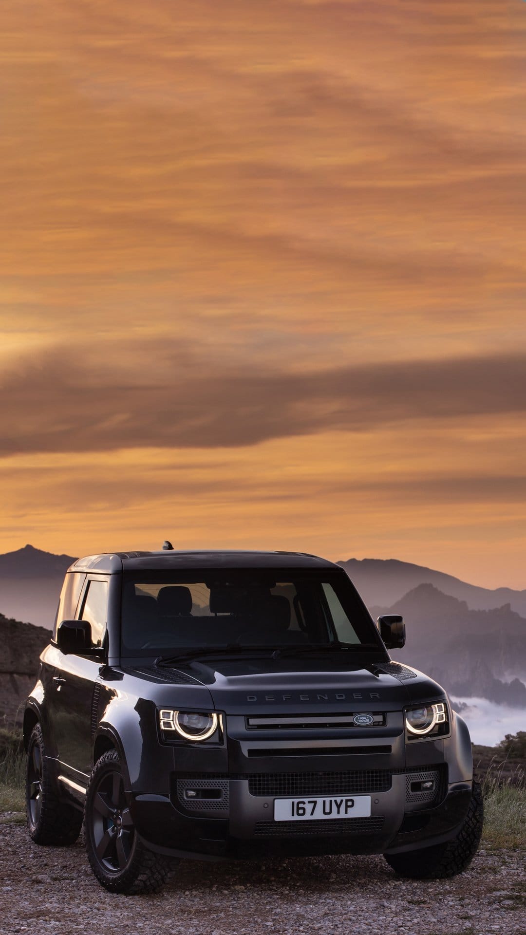 Black Land Rover Defender 90 SUV parked on a gravel path against mountain silhouettes and an orange sunset sky.