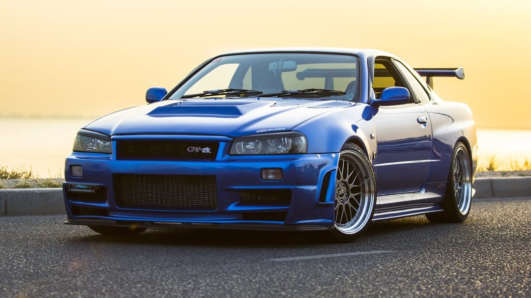 Blue Nissan Skyline GT-R R34 parked on a road at sunset with golden light and BBS wheels.