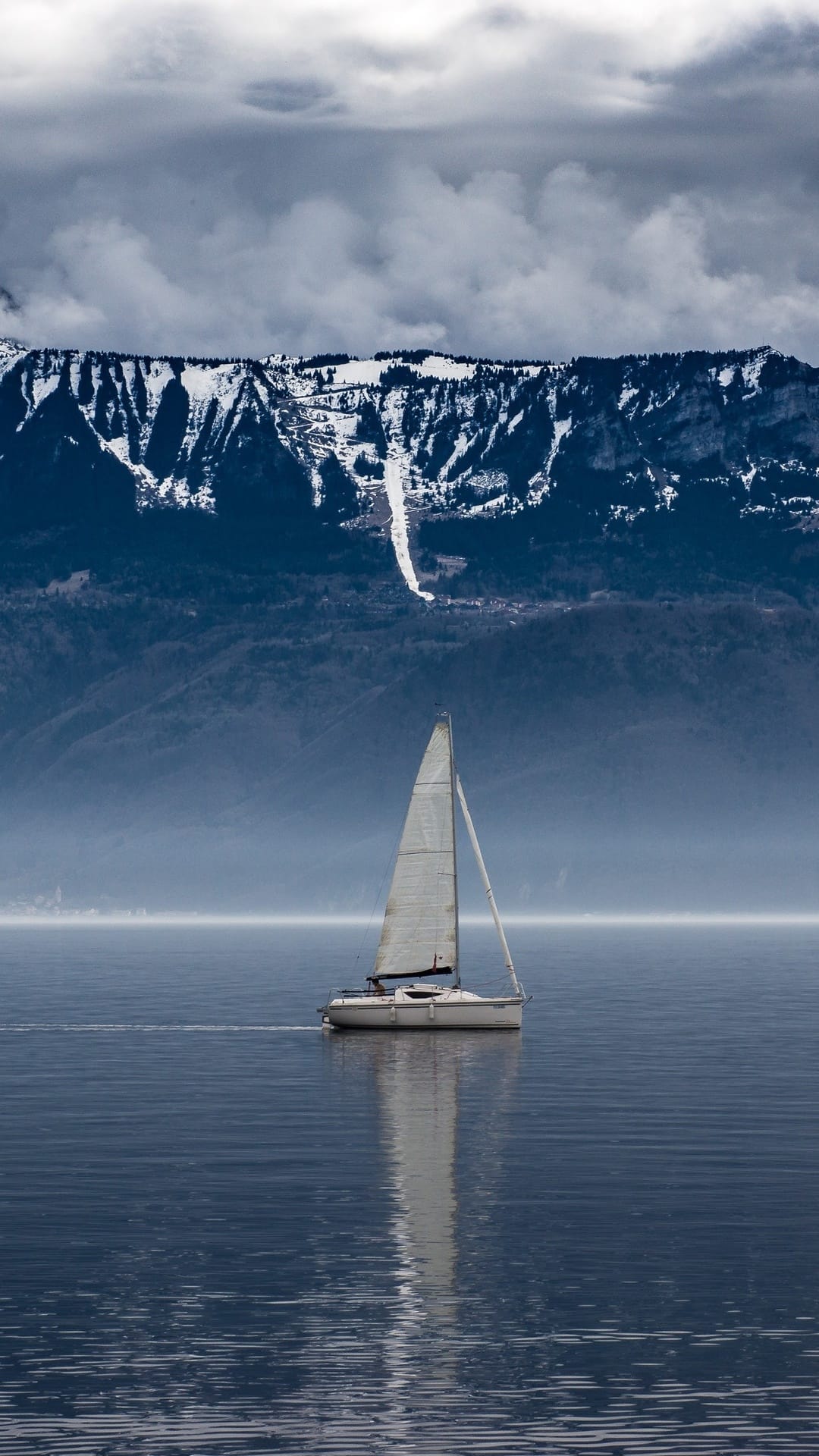 White sailboat drifts on a glassy mountain lake under snow-capped peaks and overcast skies in a vertical view.