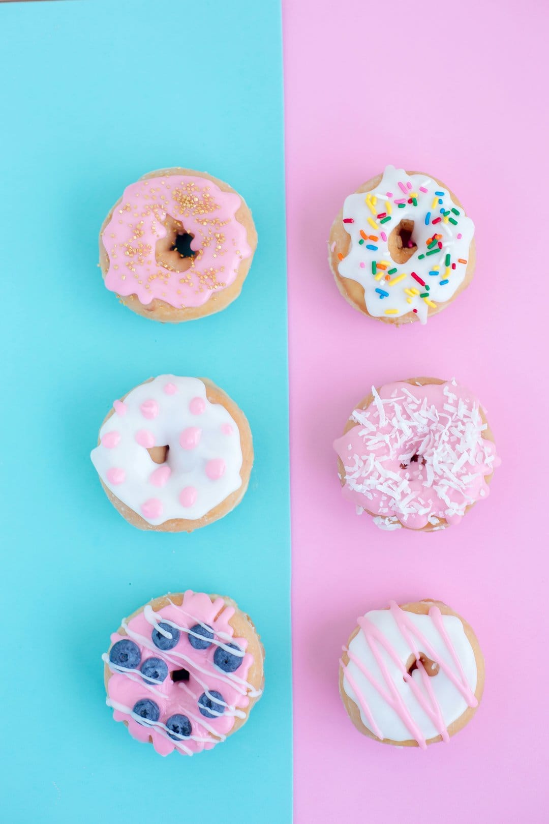 Glazed donuts with colorful sprinkles and blueberries arranged on a split pink and blue pastel background.