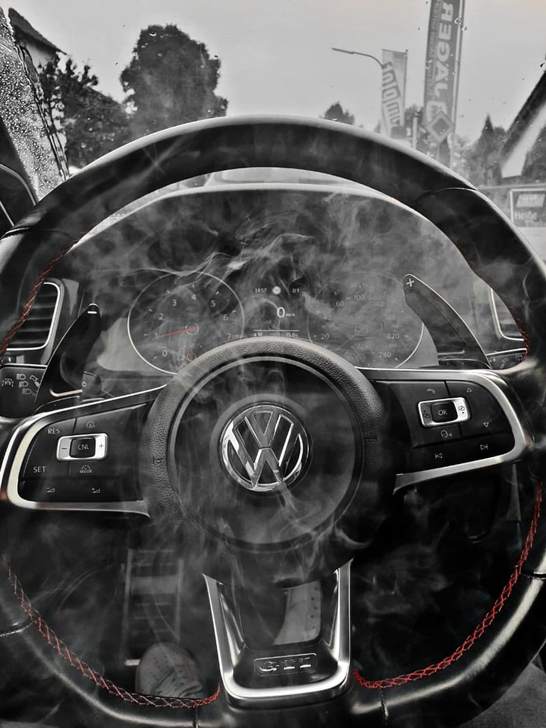 Volkswagen leather steering wheel with red stitching surrounded by smoke in front of a digital car dashboard.
