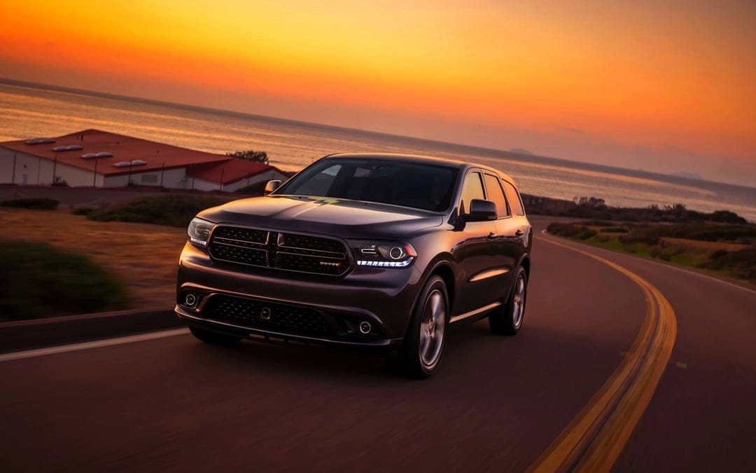 Dark metallic SUV driving on a winding coastal road during a vibrant orange sunset with calm ocean views.