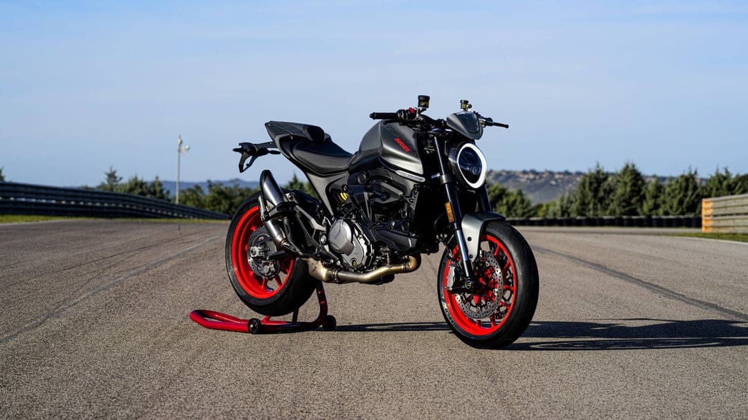 Matte grey Ducati Monster with red wheels on a paddock stand at a sunny racetrack under a clear blue sky.