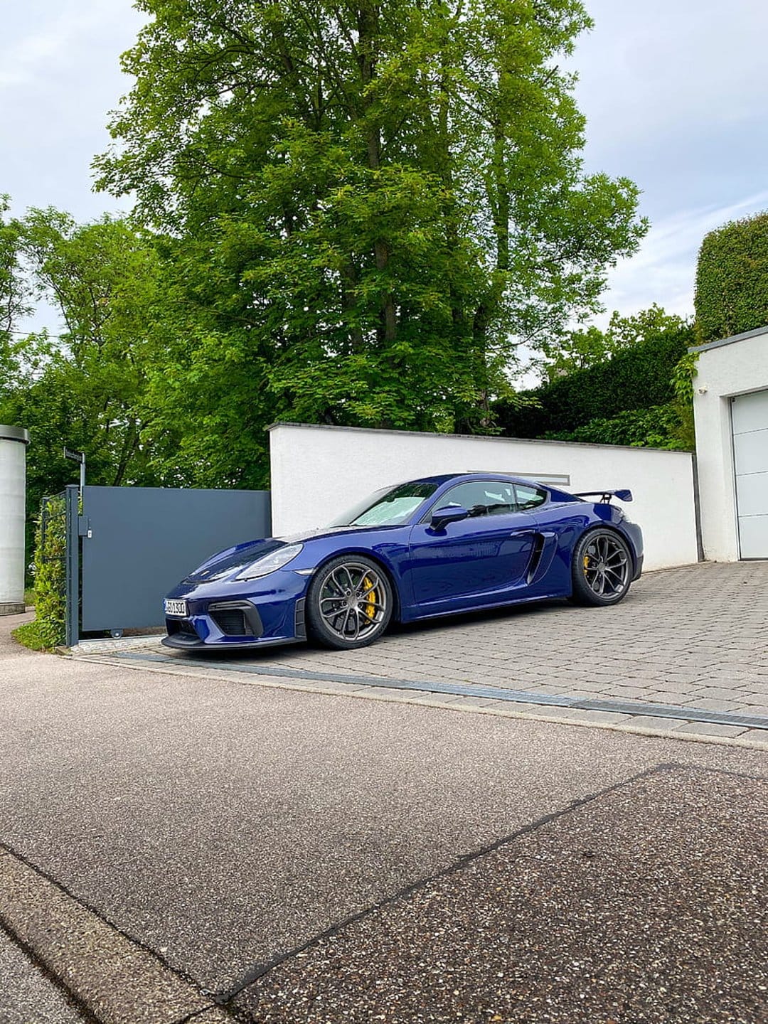 Blue Porsche Cayman GT4 parked on a stone driveway next to a minimalist white house with surrounding green trees.