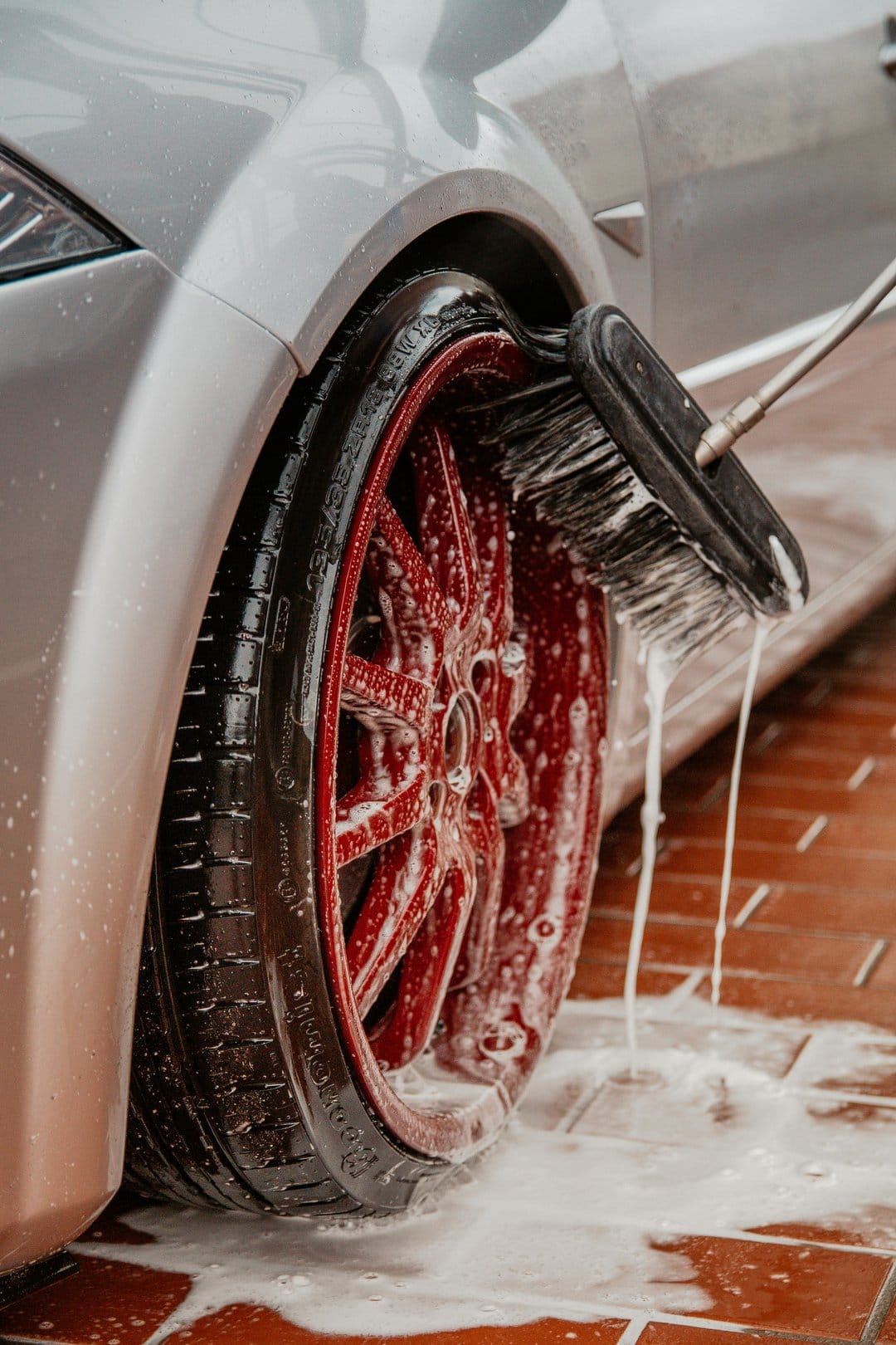 Red alloy car wheel being scrubbed with a black detailing brush as white soap suds drip onto the wet tire.