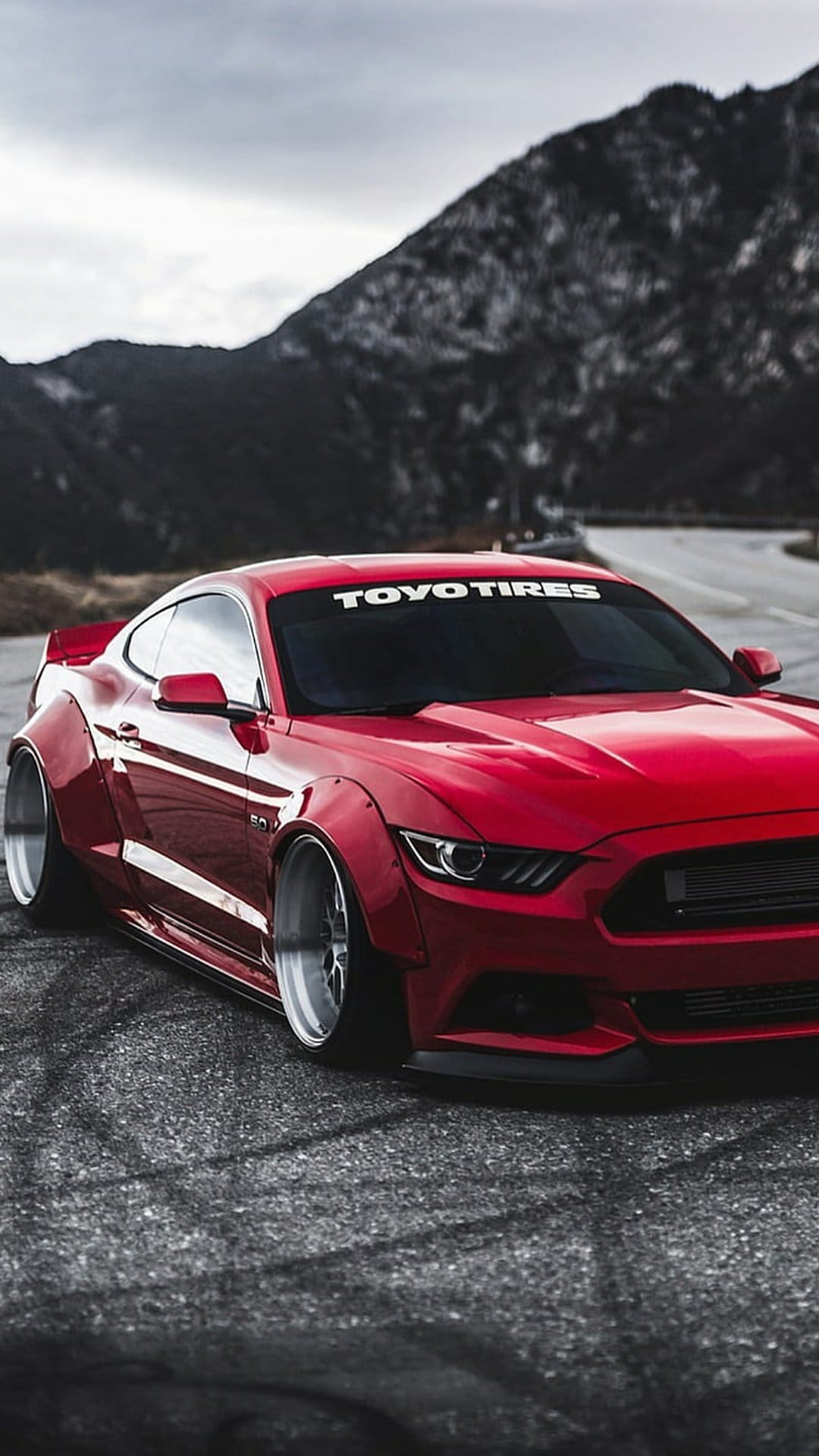 Red Ford Mustang with widebody kit and chrome wheels parked on a street with mountain peaks in the background.