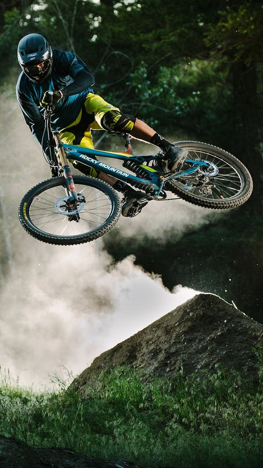 Mountain biker performs a high-air dirt jump while trailing dust against a backdrop of green forest trees.