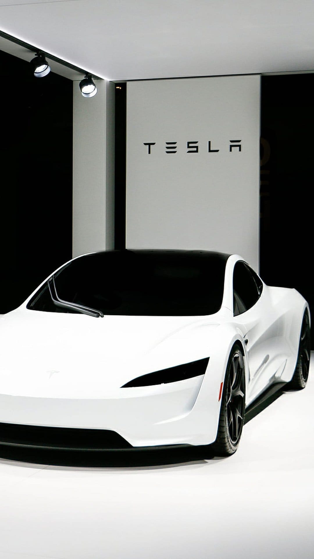 White Tesla sports car with a black roof parked in a showroom under spotlights in front of a branded backdrop.