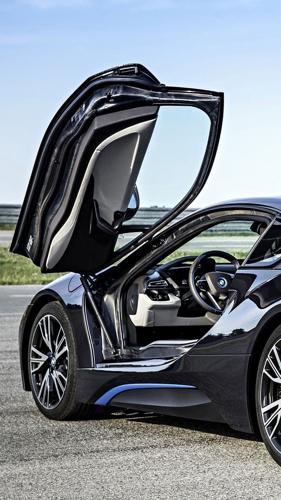 Black BMW i8 sports car with butterfly doors open on an asphalt road under a clear blue sky in bright daylight.