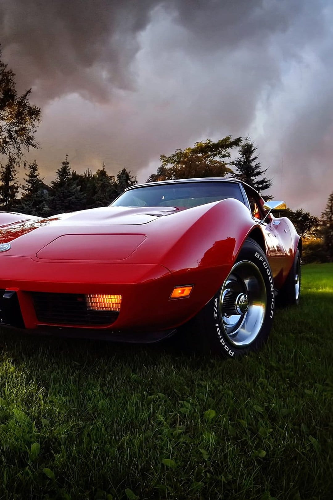 Red classic Corvette with glowing headlights parked in a green field under a dark and moody stormy sky.