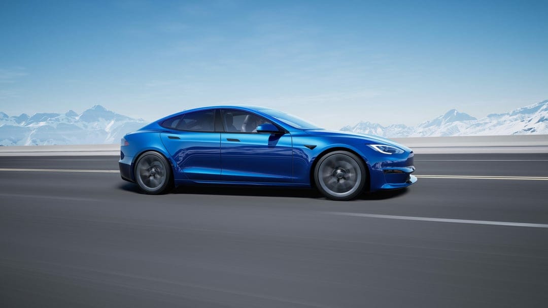 Blue Tesla electric car speeds down a wide mountain road with snow-covered peaks under a clear blue sky.