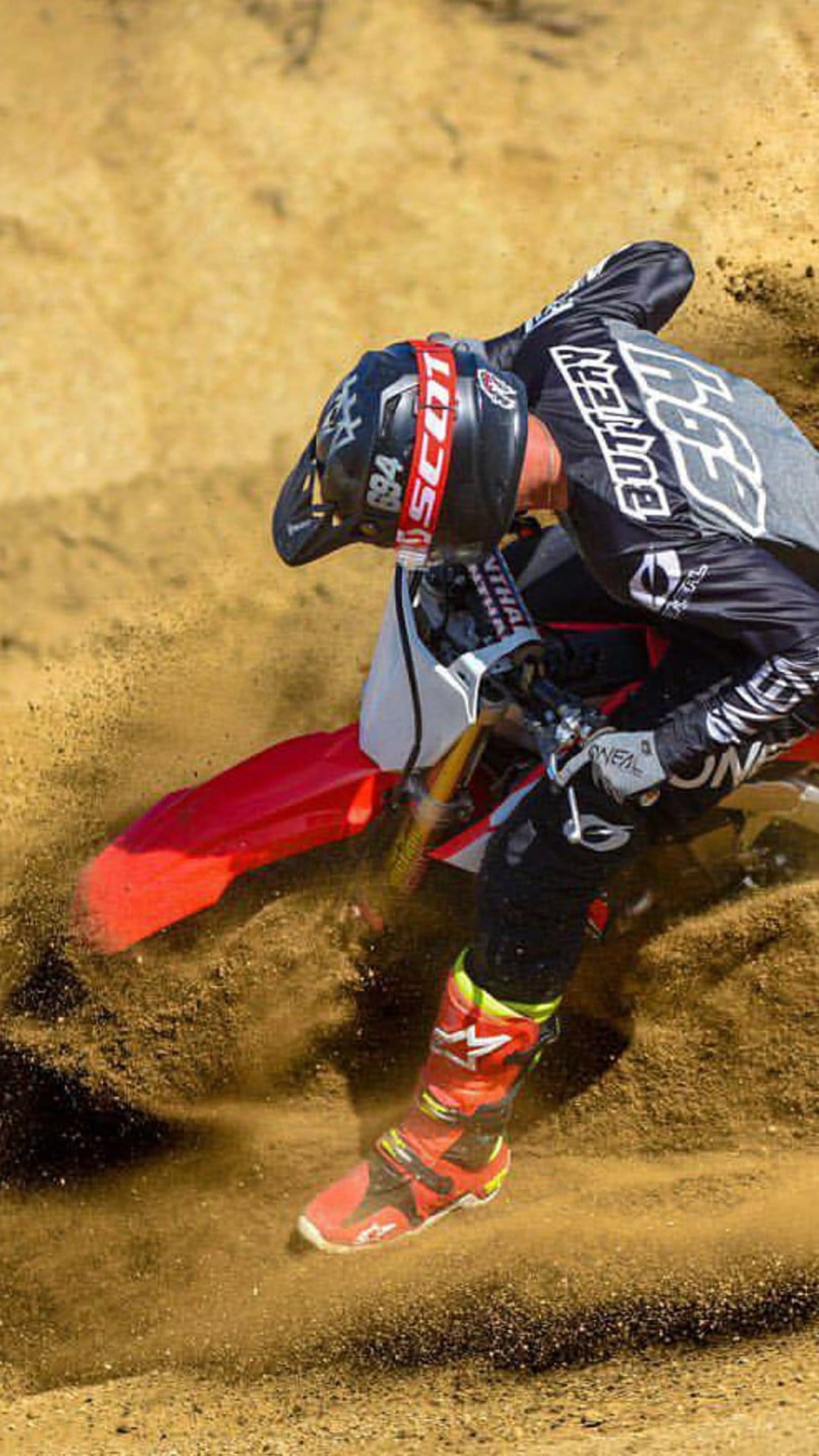 Motocross rider leaning into a sandy turn with a massive spray of dirt, wearing full racing gear and helmet.