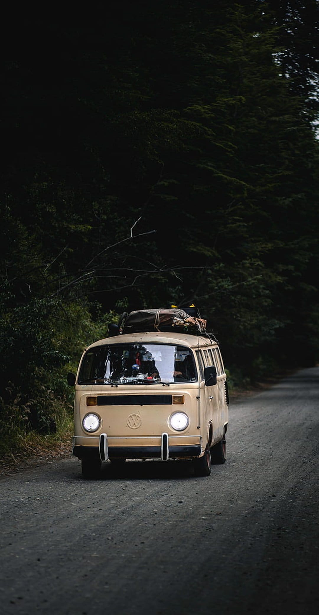 Beige vintage van with roof luggage travels a gravel road through a dense forest with illuminated headlights.