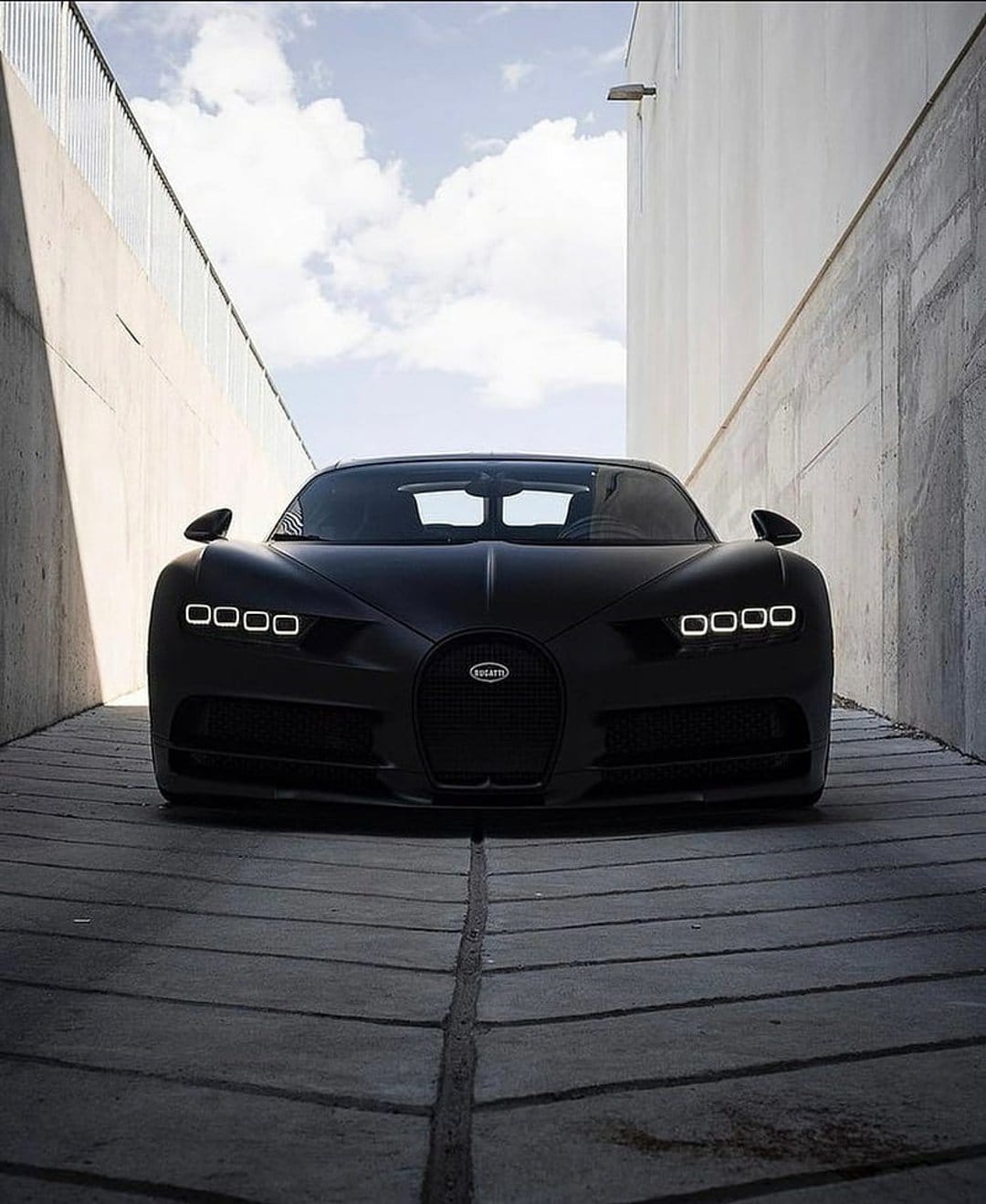 Black Bugatti Chiron with glowing headlights parked in a narrow concrete alleyway under moody evening lighting.