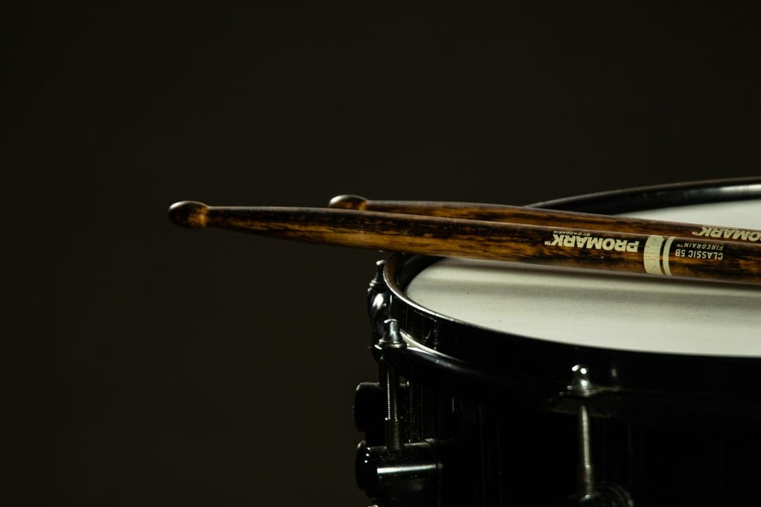 Wooden drumsticks resting diagonally on a white snare drum head with a dark, low-key lighting atmosphere.