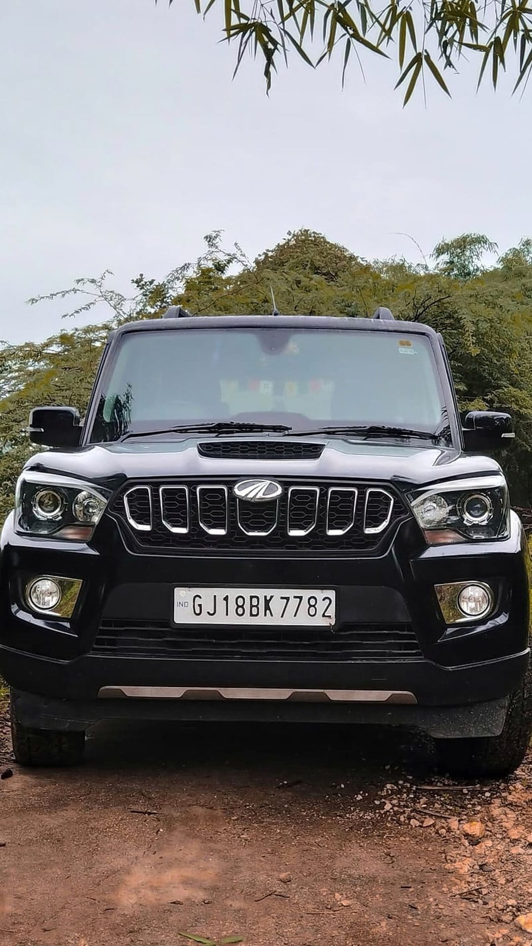 Black Mahindra Scorpio SUV with chrome grille on a dirt path surrounded by lush green trees and foliage.