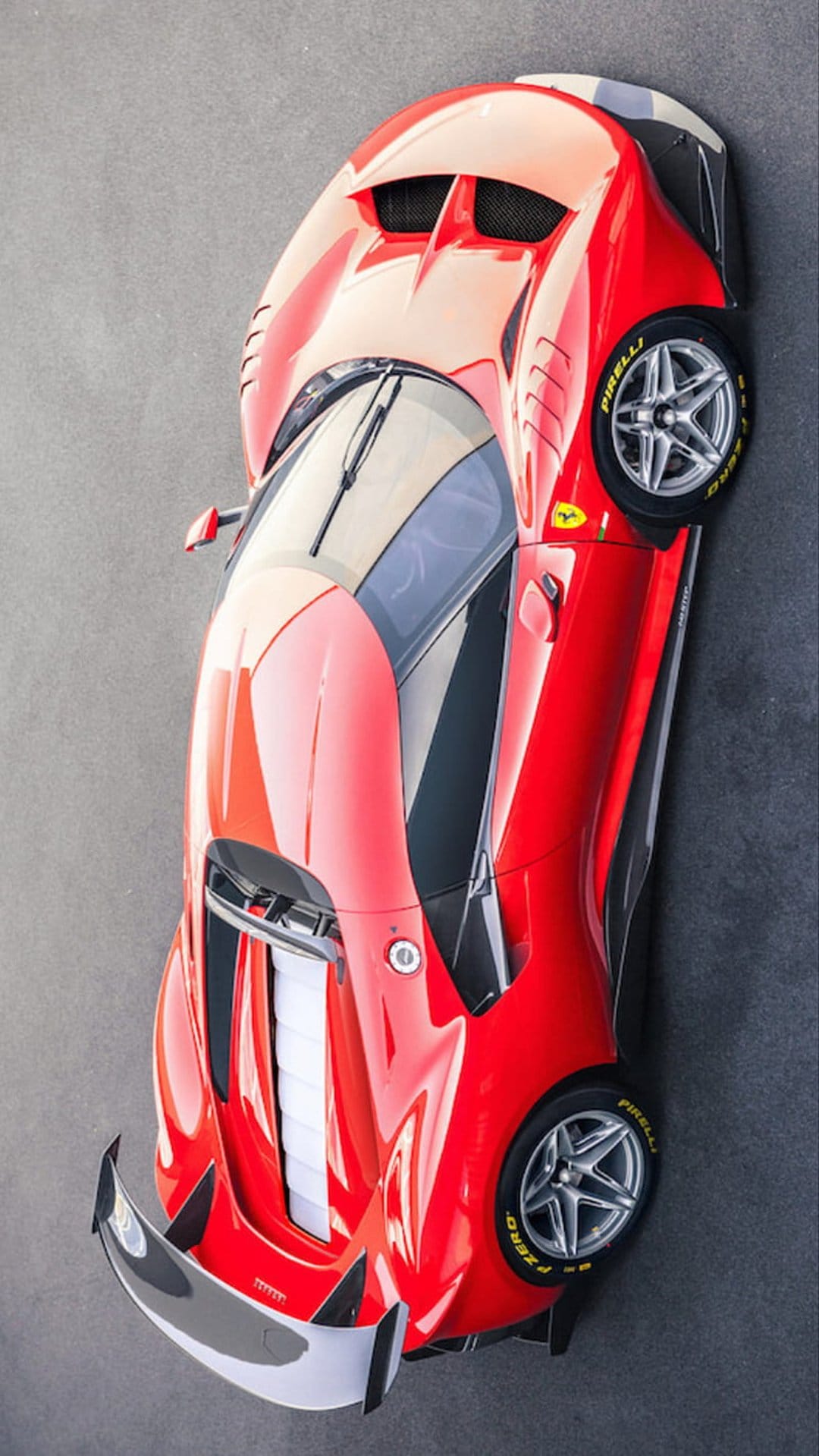 Red Ferrari supercar shot from above on a gray textured surface, showing aerodynamic vents and alloy wheels.