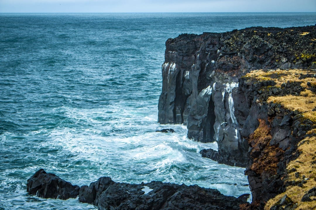 Dark basalt cliffs rise above a churning turquoise sea with white foam spraying against rugged volcanic rock faces.