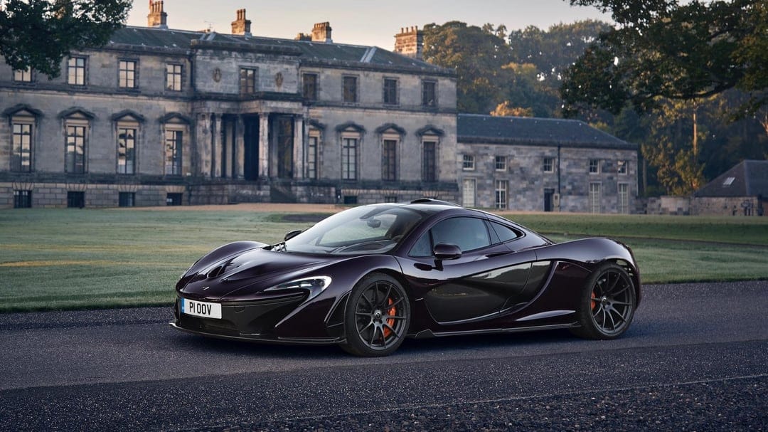 Dark purple McLaren P1 supercar parked on a path in front of a grand stone manor house during golden hour.