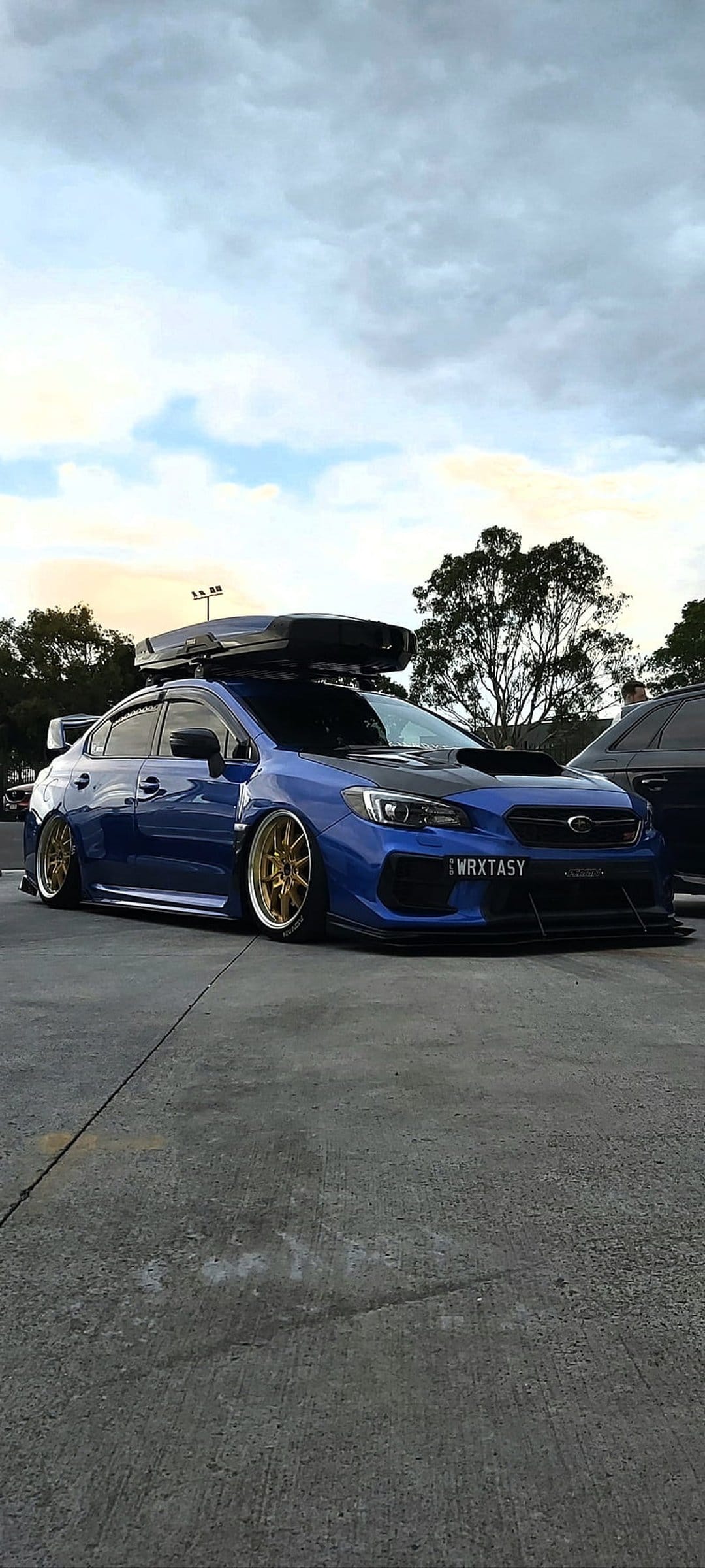 Blue Subaru WRX STI sedan featuring a black roof box and gold wheels parked on concrete under a cloudy sky.