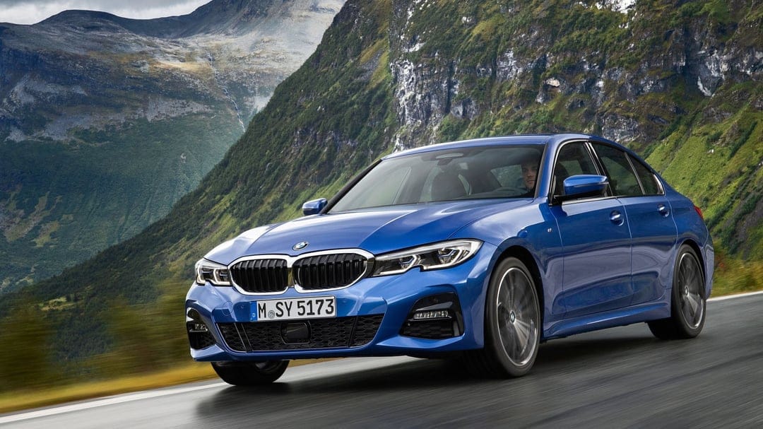 Blue BMW sedan driving on a scenic mountain road with steep green slopes and grey rocky peaks in the background.