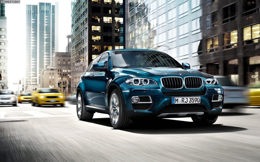 Teal BMW X6 luxury SUV driving on a city street with motion blur and tall glass skyscrapers in the background.
