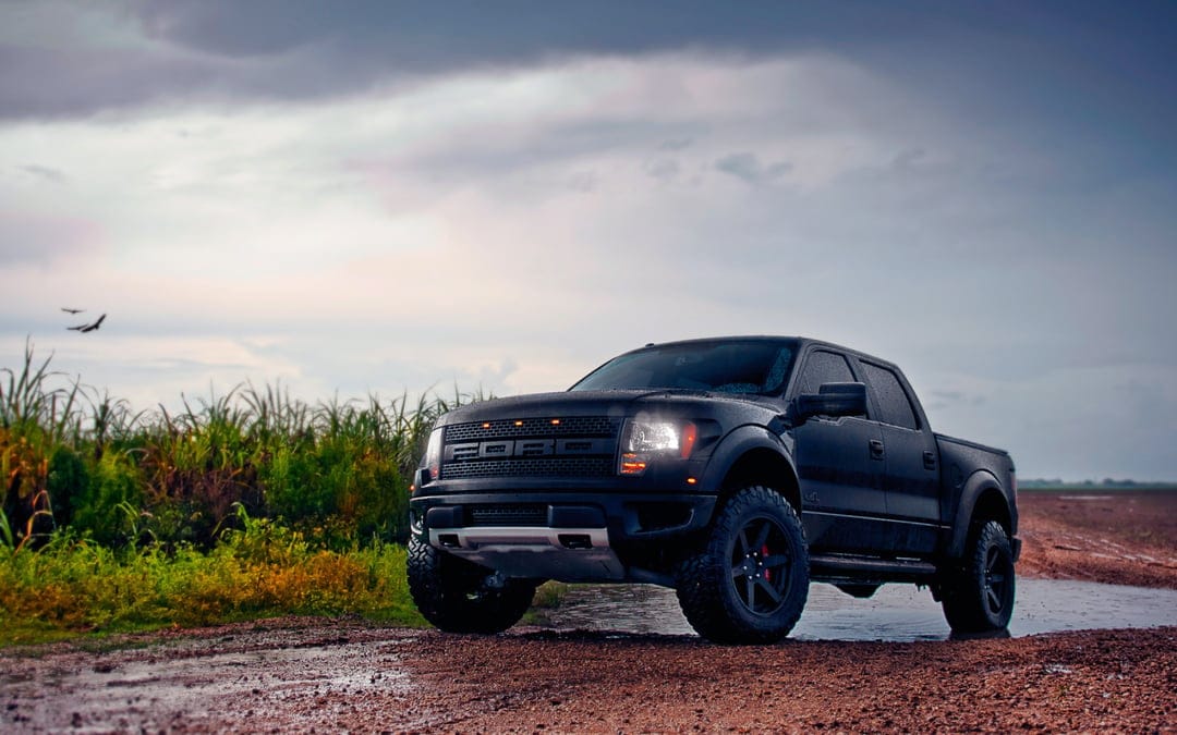 Black Ford Raptor pickup truck on a muddy road under a cloudy sky with headlights and amber grille lights on.