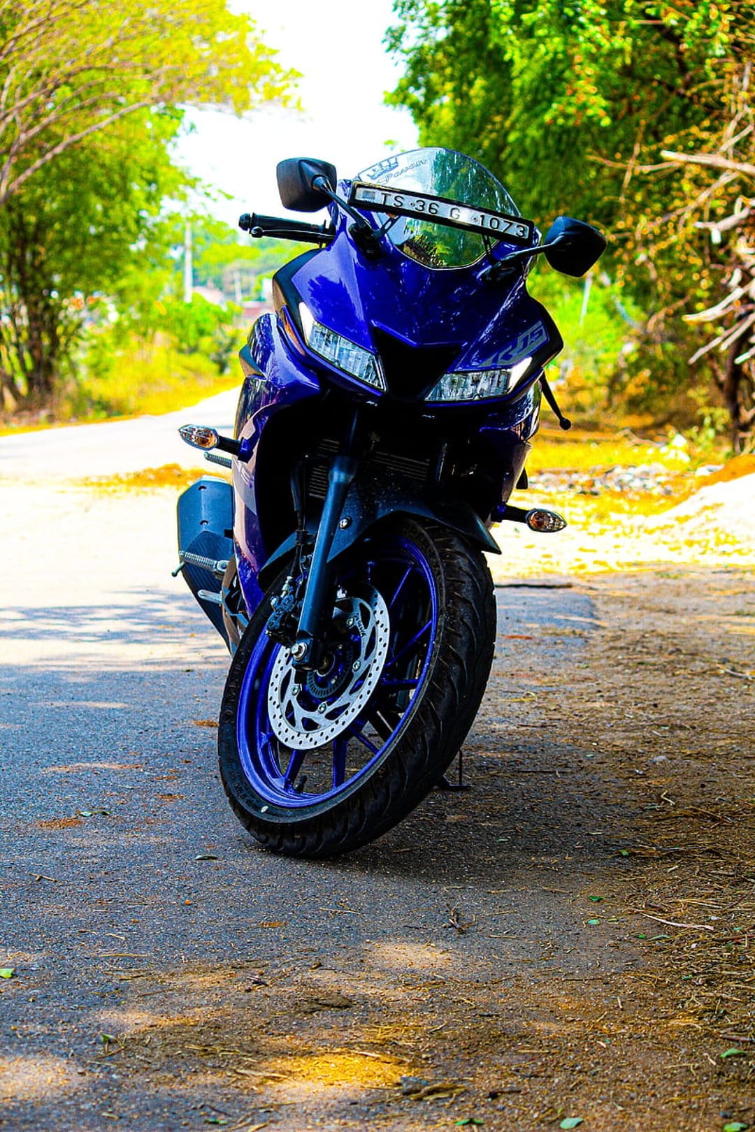 Yamaha R15 V3 motorcycle in metallic blue with LED headlights parked on an asphalt road near green trees.