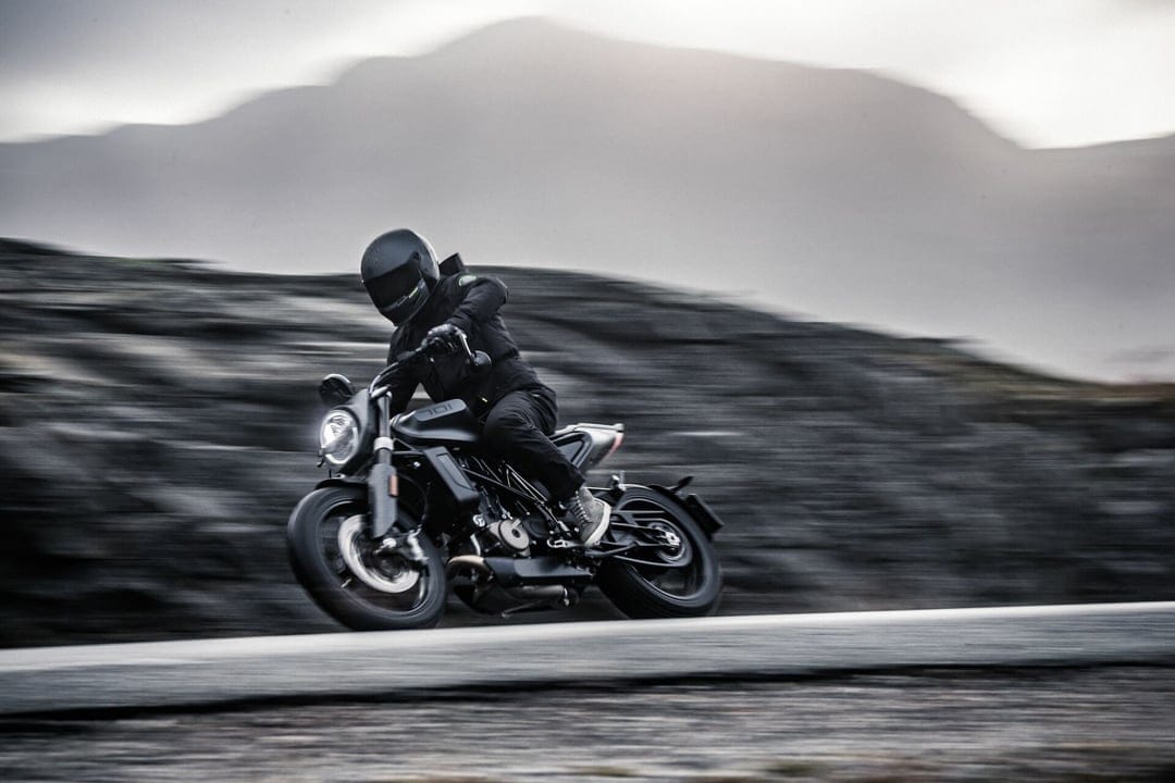 A motorcyclist in black gear riding a dark street bike on a paved road with mountain scenery and motion blur.