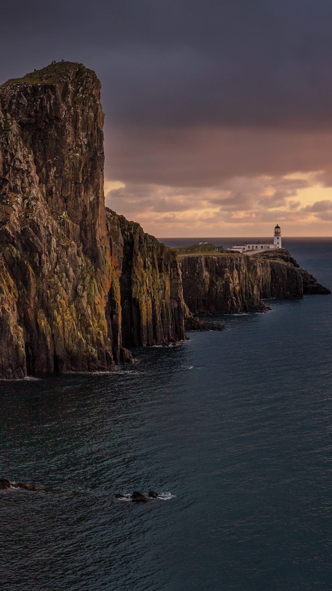 Neist Point Lighthouse perched on steep cliffs overlooking the dark Atlantic Ocean under a stormy twilight sky.