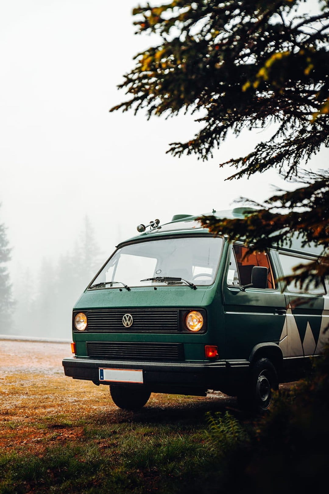 Green vintage Volkswagen van parked on grass surrounded by tall trees and thick white fog in a secluded forest.