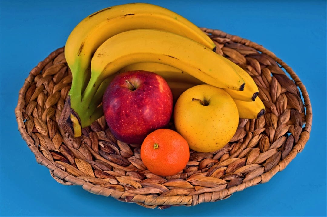 Ripe bananas, red and yellow apples, and a bright orange sit inside a woven wicker basket on a flat surface.
